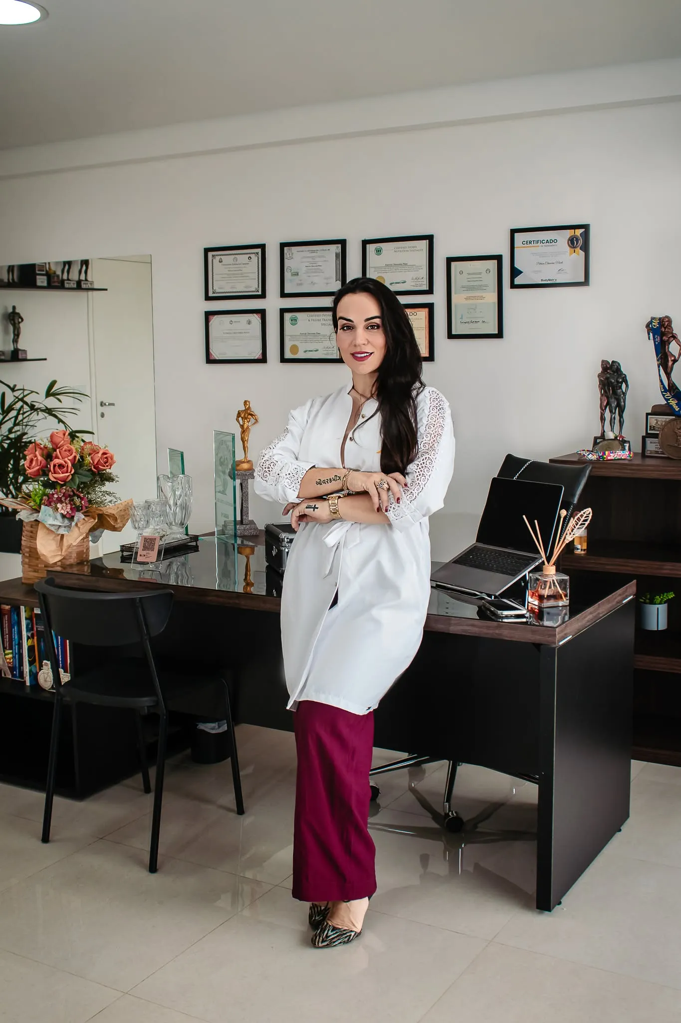 Patrícia Chiaverini de jaleco no consultório, com diplomas na parede e troféus na estante